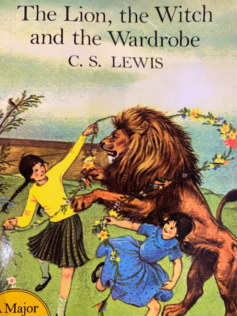 Book cover of "The Lion, the Witch, and the Wardrobe" written by C.S. Lewis. Aslan the lion dancing in a field with Susan and Lucy