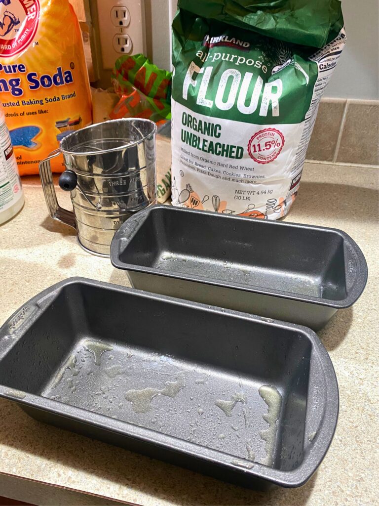 Two greased loaf pans with a flour sifter and coconut oil in the background