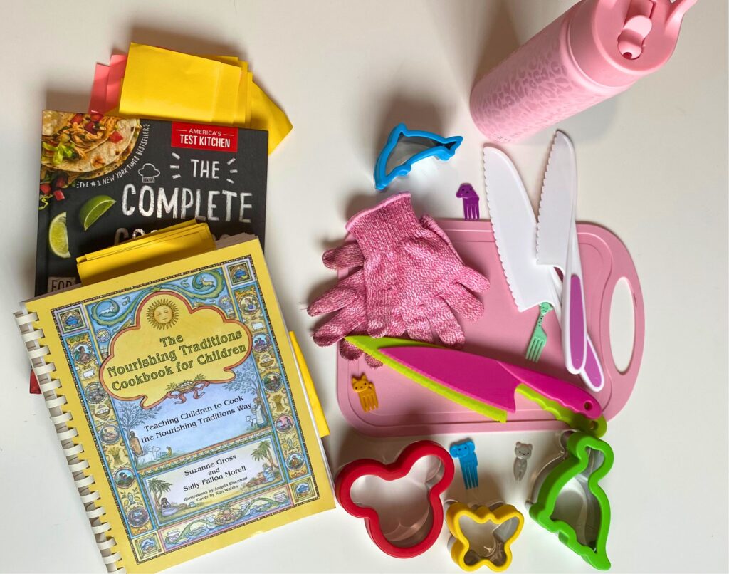 Kids kitchen products from far away: America's test kitchen kids' cookbook, Nourishing traditions cookbook for children, sandwich cutters, food picks, stainless steel water bottle (pink cheetah print from Simple Modern), and plastic knife and glove set on top of a pink cutting board