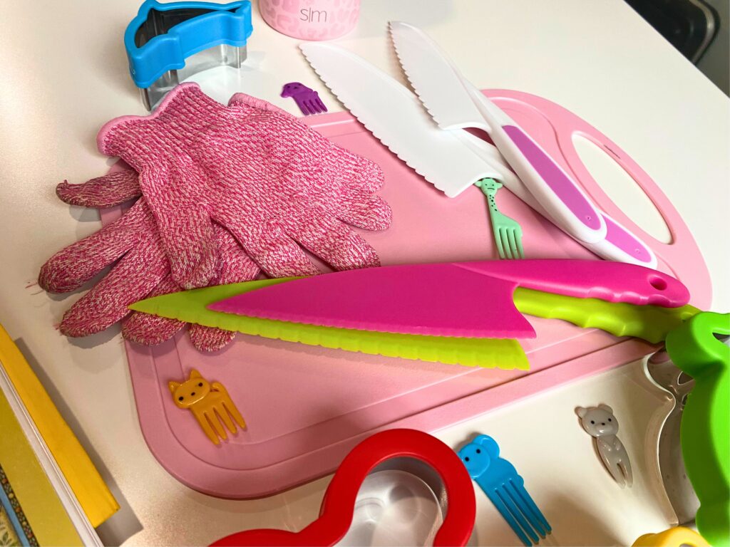 pink kids kitchen safety gloves and four plastic knives on top of a pink cutting board surrounded by food picks and sandwich cutters