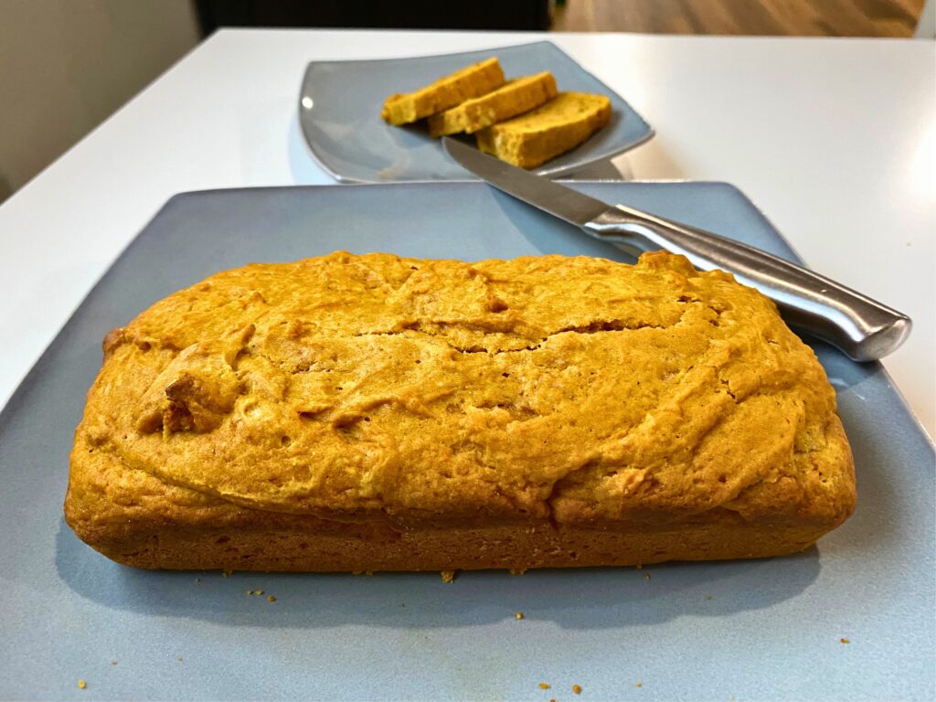 A full pumpkin bread loaf uncut plated on a grey plate with a knife in the foreground and three pumpkin bread slices in the background