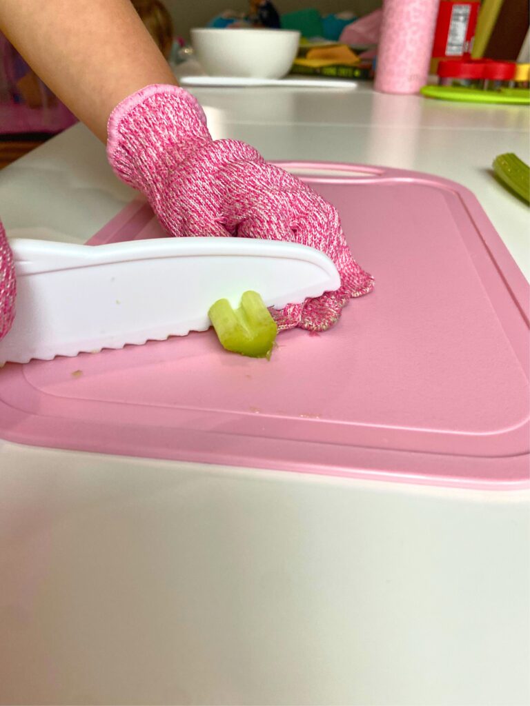 Young child using plastic knife and pink kitchen safety gloves to cut celery on a pink cutting board