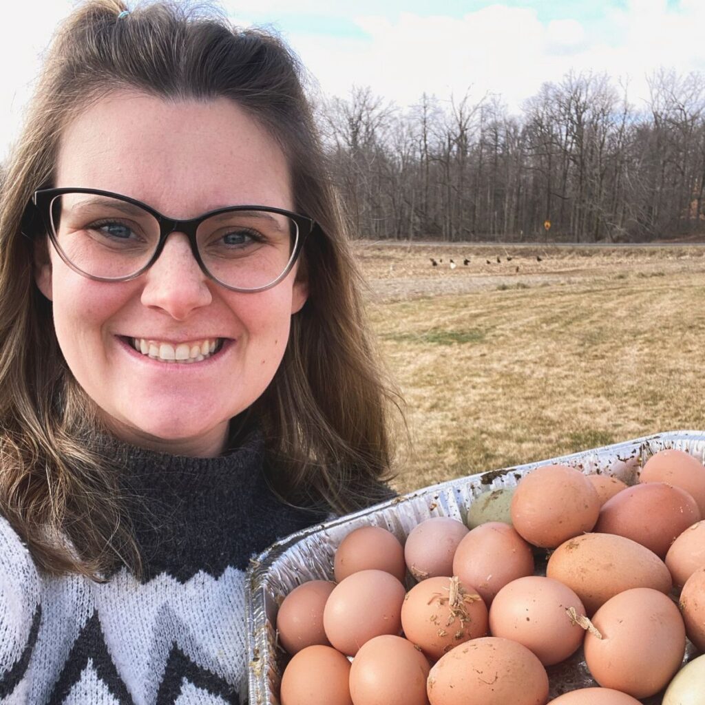 Woman smiling outside by free ranging chickens in a corn field. She is holding up a metal tray filled with twenty colored eggs, brown and green.