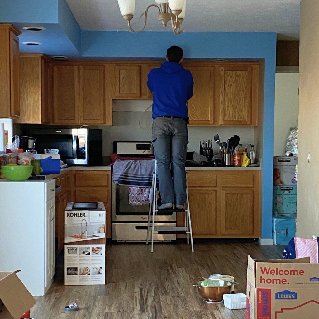 Man standing on a step stool in a messy kitchen working on a microwave above the oven