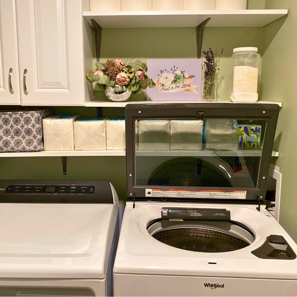 A laundry room with a washer on the right opened and a shelf above with dried flowers as decoration and homemade laundry detergent in a recycled container to the right