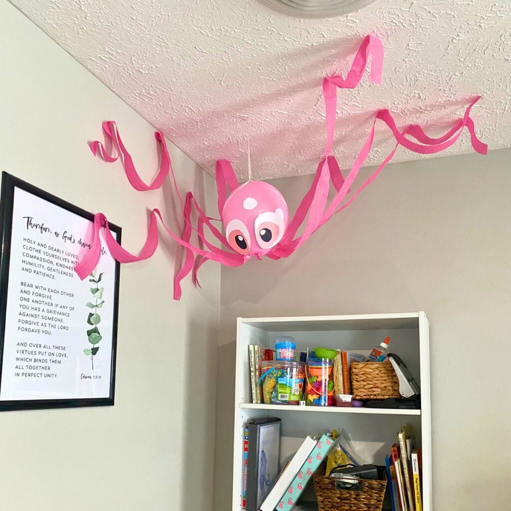 A balloon and streamers in the corner of where a wall and the ceiling meet in the shape of a pink octopus