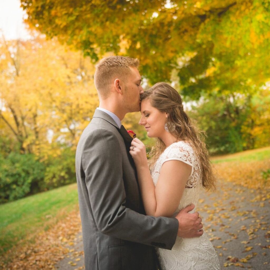 Man and woman on their wedding day outside. Man is kissing woman on the forehead as her eyes are closed and she grips his lapel.