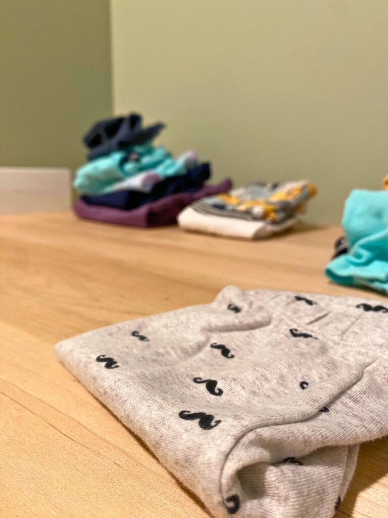 Newborn boy's grey pants with black mustaches on them folded on a wooden countertop with piles of clothes for a baby boy in the background