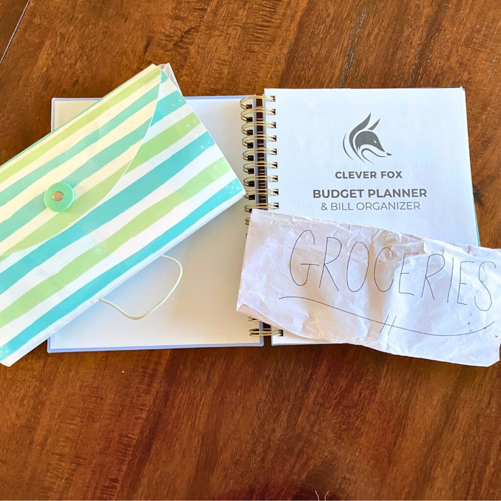 Blue, green, and white striped accordion file, budget planner & bill organizer, and an envelope for the cash envelope budgeting system labelled "Groceries" on a tabletop