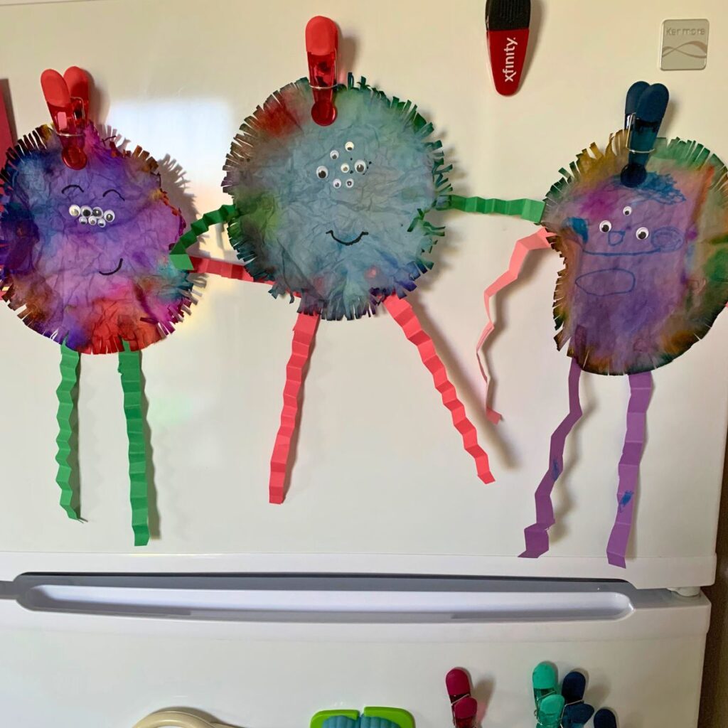 Three monsters with googly eyes made from colored coffee filters craft hung on a fridge