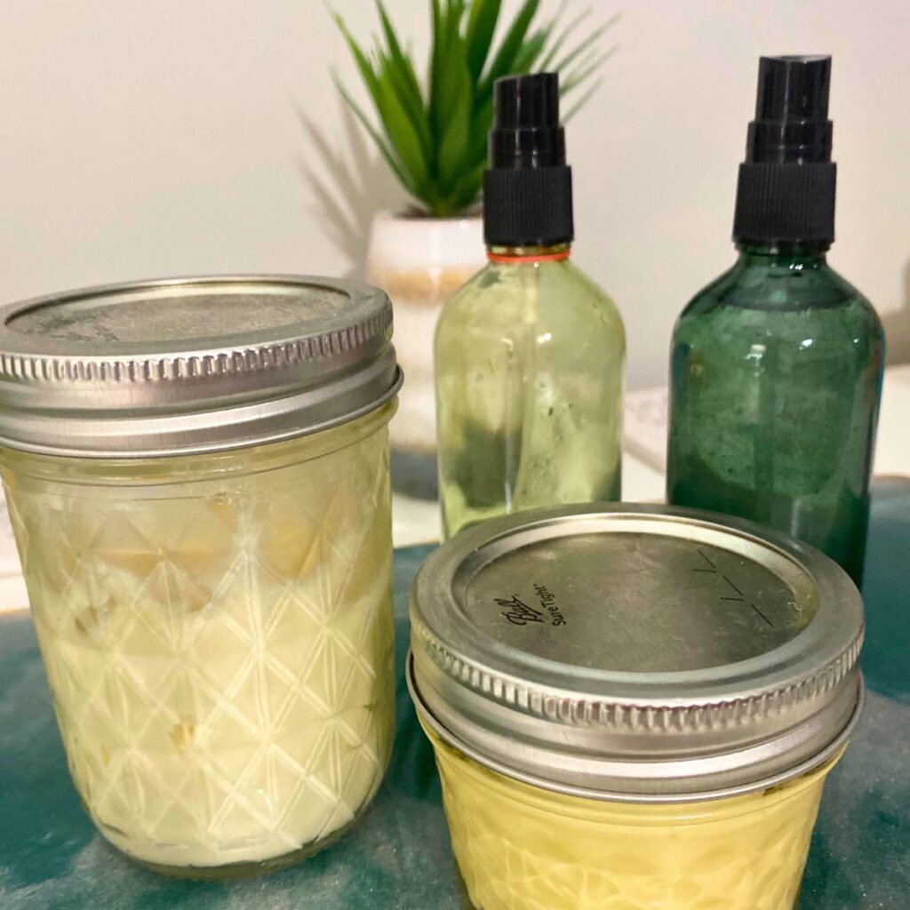 Two glass spray bottles filled with essential oils and water as a perfume substitute behind two mason jars filled with homemade lotion scented with essential oils