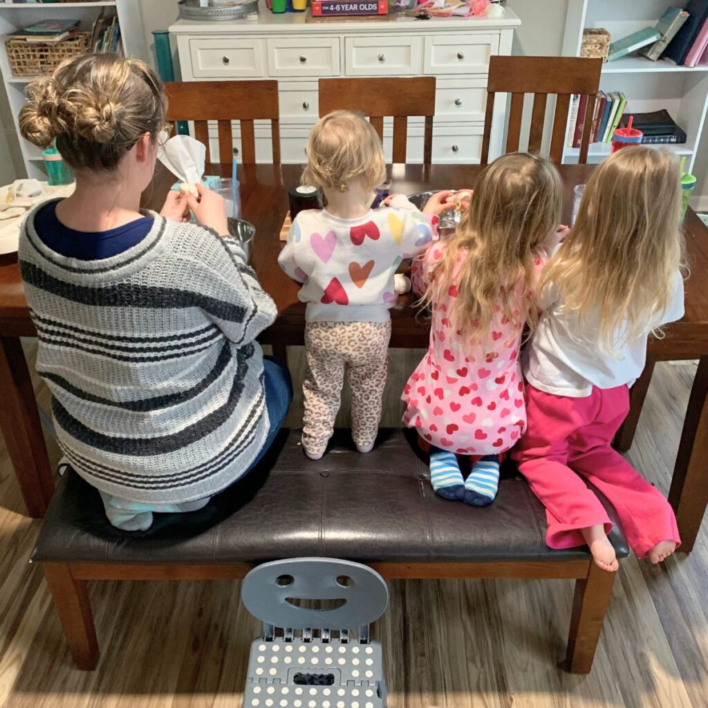 Woman and three young children sitting on a bench at the kitchen table peeling garlic (taken from the back)