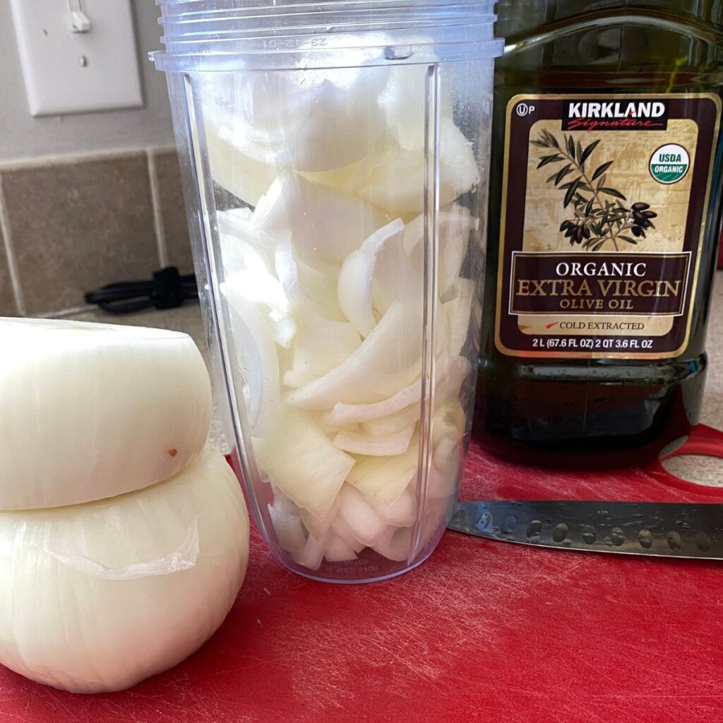 Two peeled onions stacked on one another next to a Nutribullet blender cup full of roughly chopped onion pieces. To the very right is a large bottle of organic extra virgin olive oil and a knife