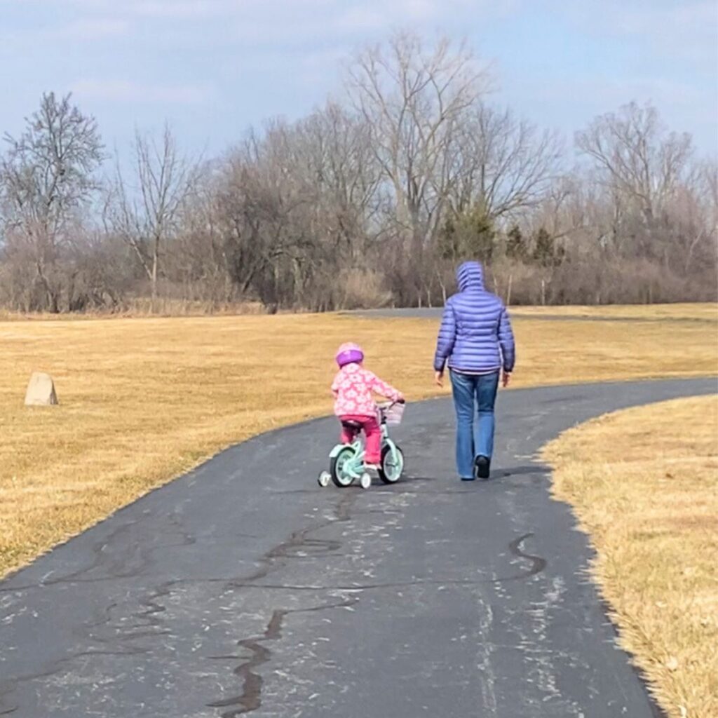 Toddler in a pink floral jacket wearing a helmet riding a mint green bike with training wheels for the first time 