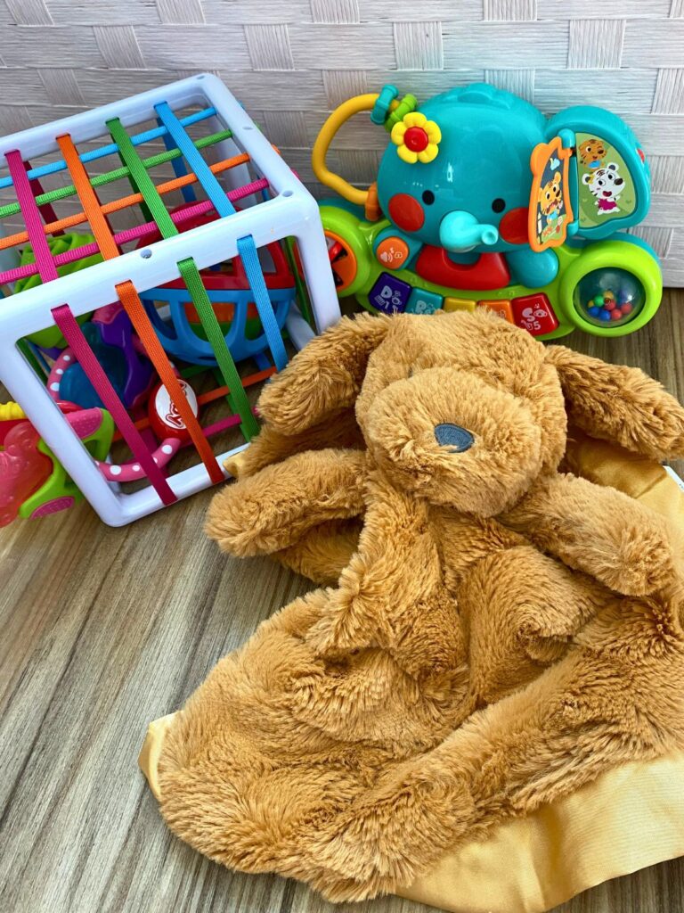 Baby's toys sitting in a pile on the floor: a stuffed dog blanket toy, a cube with toys inside, and an elephant piano