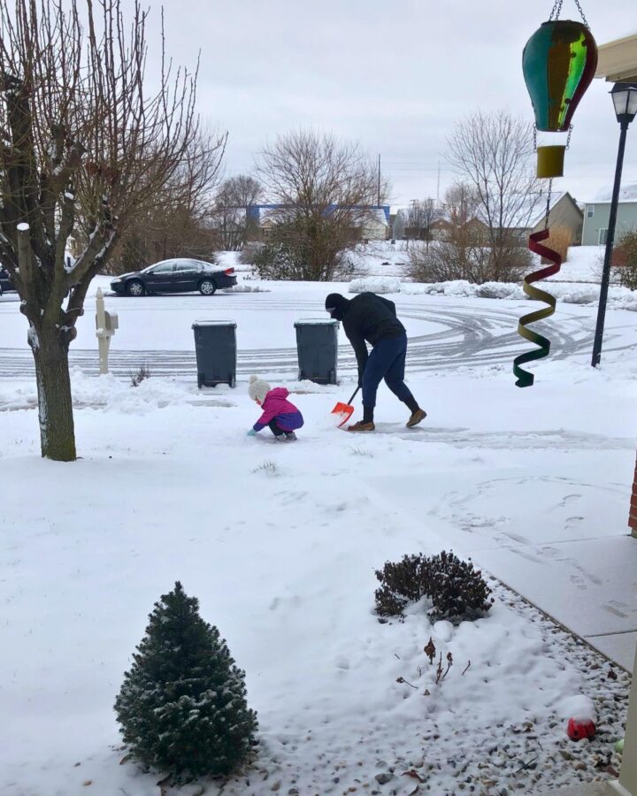 Child kneeling in the snow playing while man shovels the driveway beside her