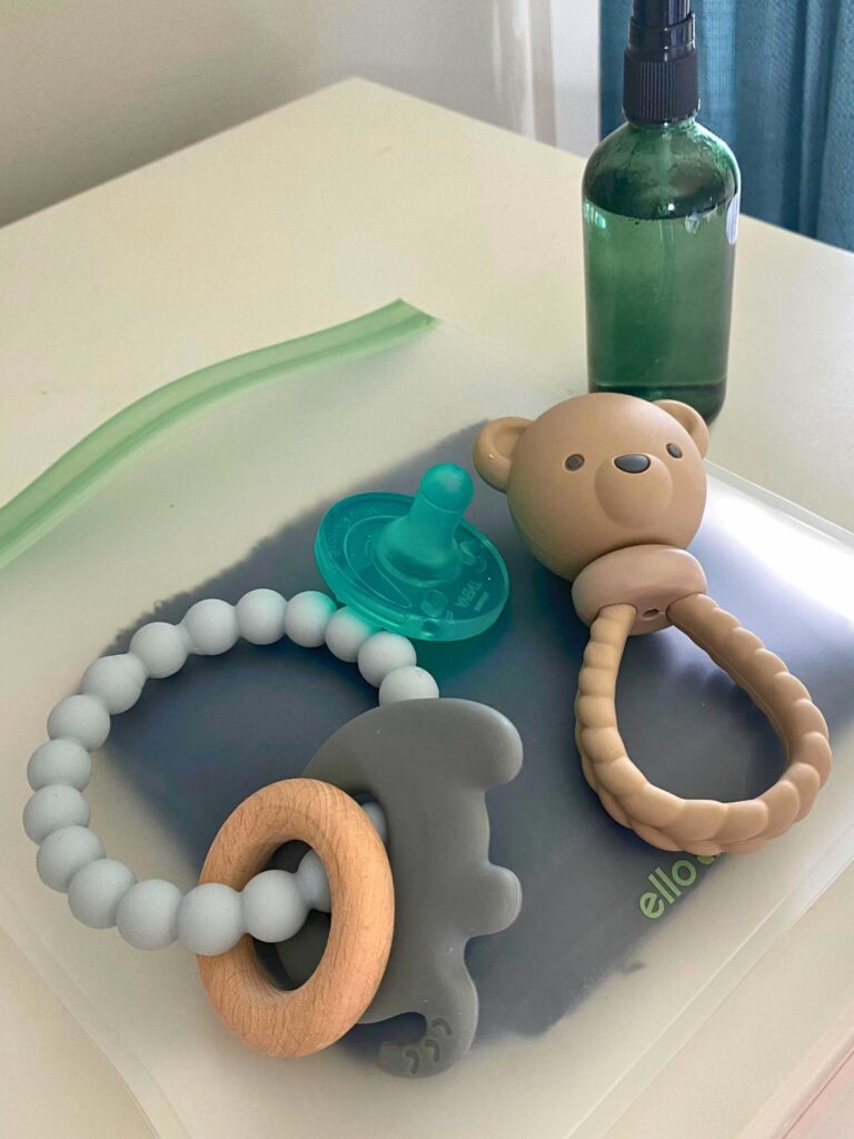 Clean silicone baby toys and pacifier on a white dresser with a glass spray bottle filled with vinegar cleaning solution in the background